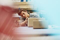 Male blue-collar worker checking parcels. Caucasian man. Checkered shirt. Smooth-shaven chin. Conveyor belt. Logistics environment. Primary color light-red. Secondary colors light- blue and cream.