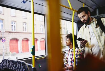 Man on his phone standing in a bus. Woman sitting down in the background.