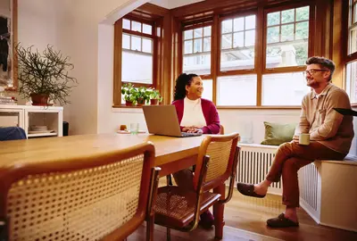 Female sitting at a table with laptop talking to male with coffee mug at home
