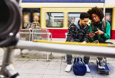 male and female looking at phone while sitting on a bench on a train platform.
