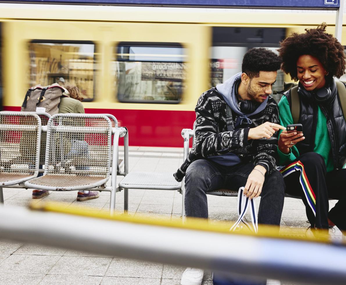 Man and woman looking at phone while sitting on a bench on a train platform.