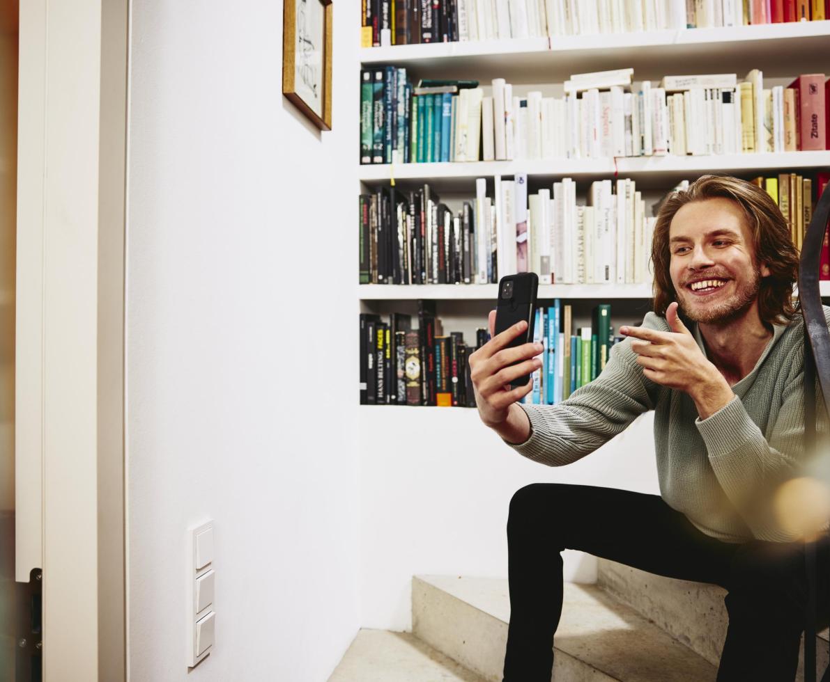 Smiling man sitting on stairs, making a selfie. bookshelf in background