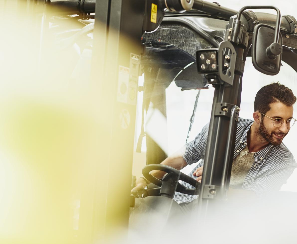 Male caucasian worker operating a forklift in a logistics environment. Looking to his left. Blue-collar. Smiling.  Wearing glasses. Groomed beard and moustache. Checkered shirt. Primary color cream.